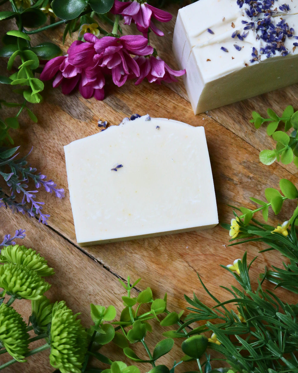 Two bars of lavender soap on a wooden surface with greenery and flowers.